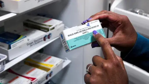 Reuters A box containing the vaccine against measles, mumps and rubella, being held by a woman with her right hand, with the index finger of the left pointing to the writing on it.