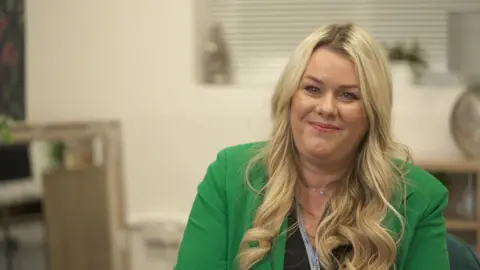 Kerry with long blonde hair in a bright green jacket sits indoors against a neutral background smiling. 