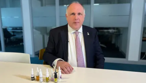 Alastair Richards is looking at the camera, wearing a suit and tie sat at a table with seven nasal spray tanners in front of him