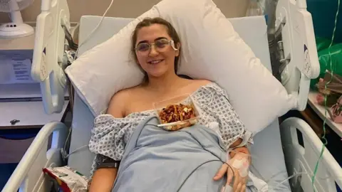 Ami Clarke Ami is propped up in a hospital bed with a large white pillow behind her head and tubes coming out of her hand - there is a pot of pasta in front of her and she is smiling