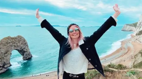 Clare Elgar Clare Elgar, who has MS, stands on a beach, smiling at the camera, arms raised in the air