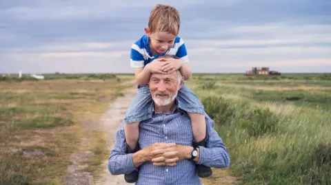 Getty Images An older man carries a young boy on his shoulders during a walk outside. The relationship is one of grandfather and grandson. 